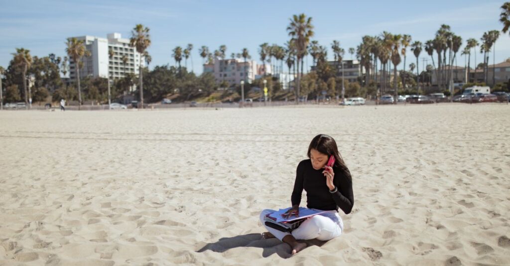 Woman on a sandy beach using a cellphone and handling paperwork, epitomizing remote work and leisure.