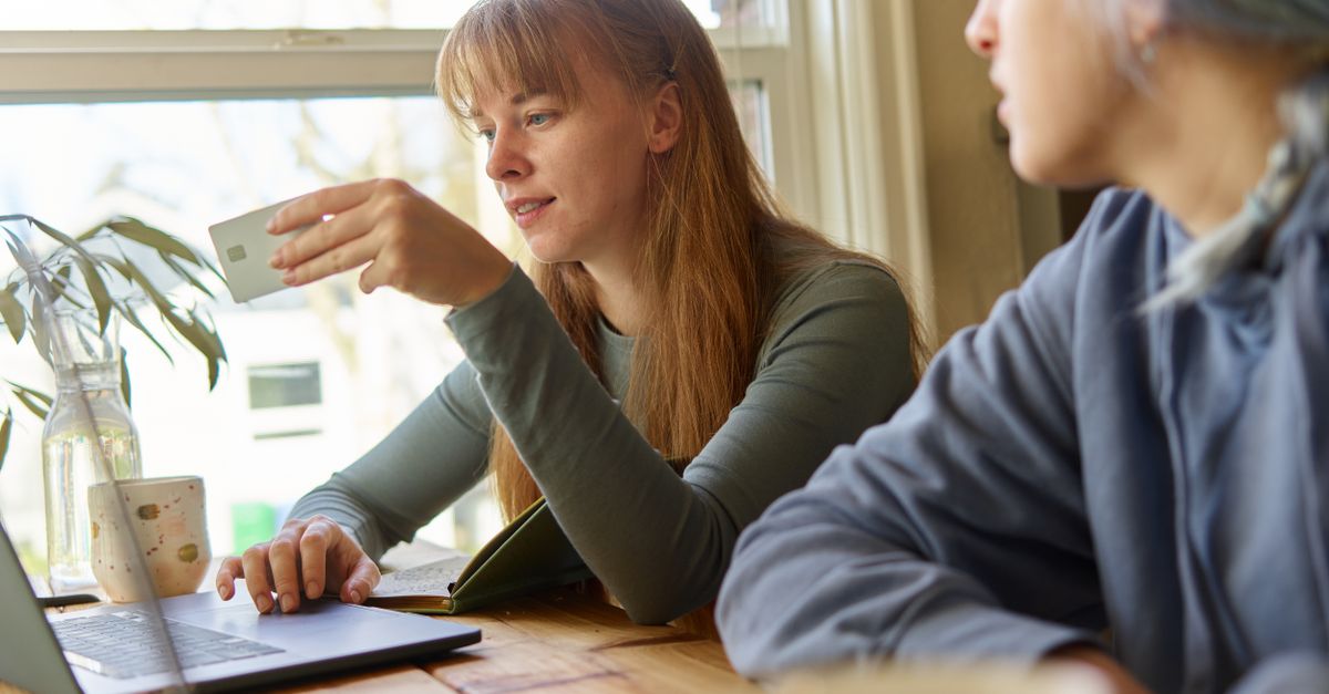 Two young women in casual attire discussing online payments using a laptop at a wooden table near a window.