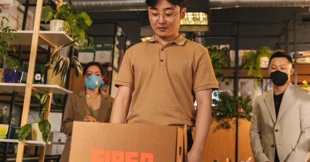 Man standing by cardboard box labeled 'fired', in an office surrounded by colleagues.