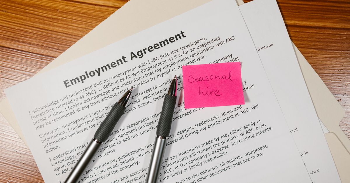 Top view of employment agreement contract with pens and seasonal hire note on wooden desk.