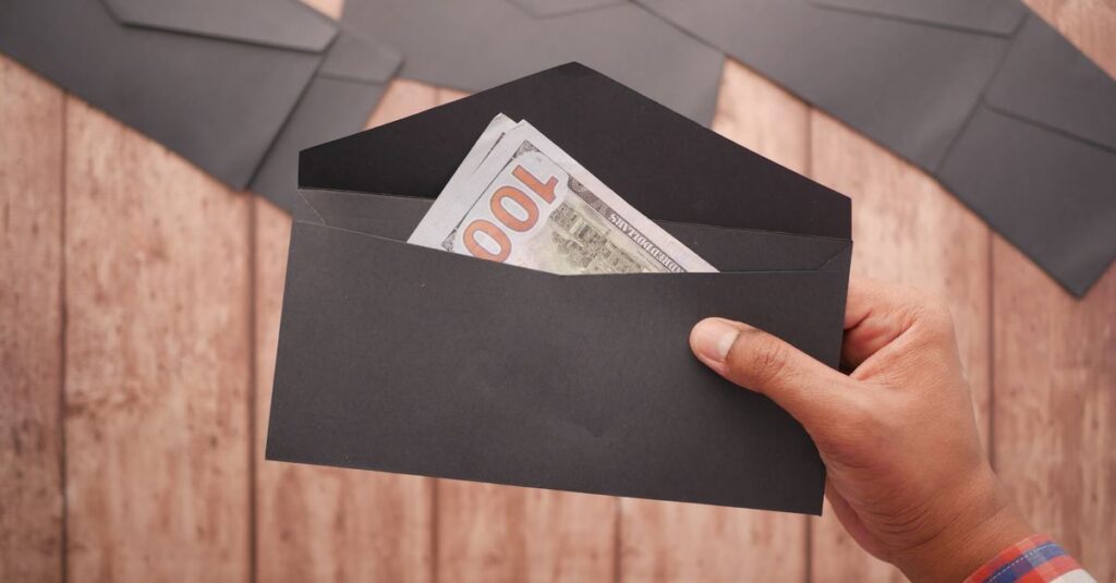 A hand holds a black envelope containing a 100 dollar bill on a wooden surface.