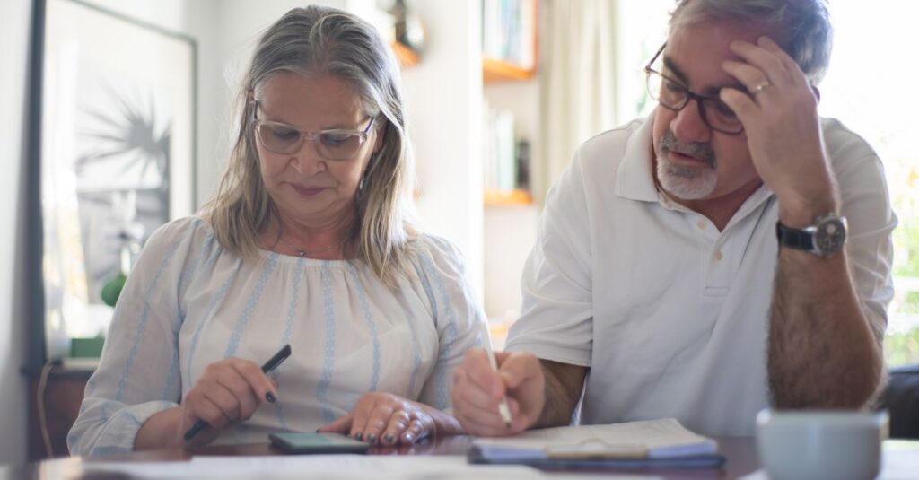 Senior couple calculating expenses at home office desk with documents and notes.