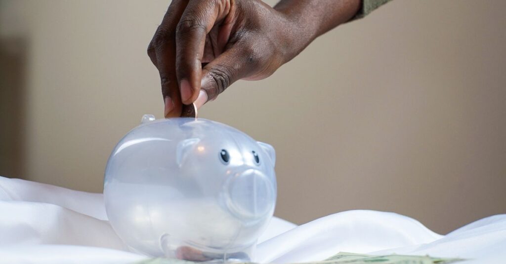 Close-up of a person's hand placing coins into a transparent piggy bank to save money.