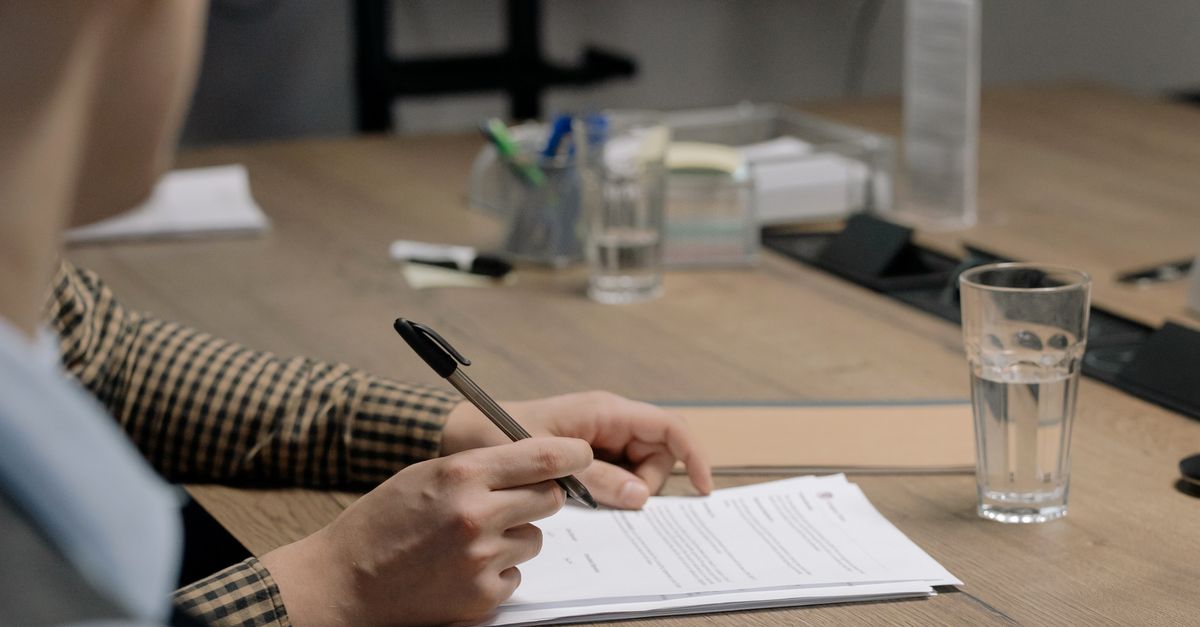 Person in business attire signing a document at a wooden table in an office setting.